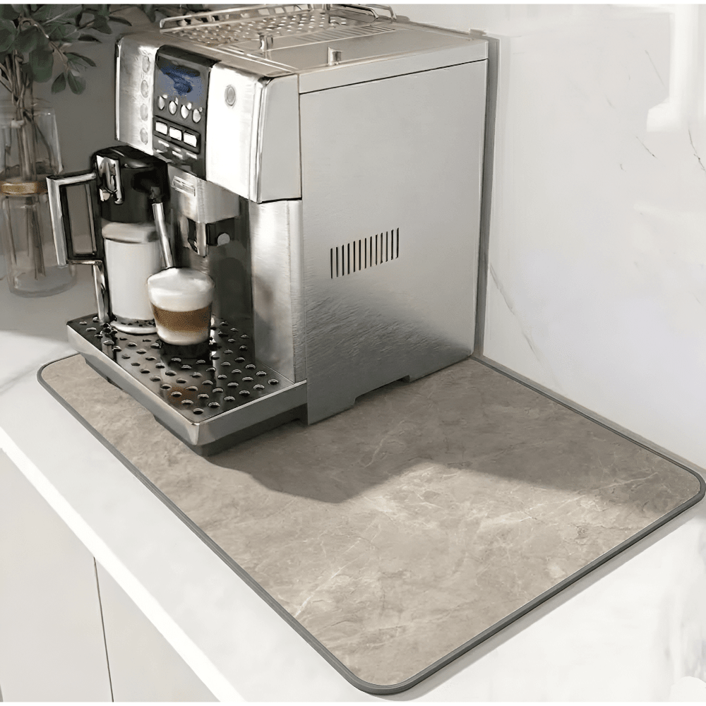 Beige matta med marmormönster och grå kant under en silverfärgad kaffemaskin på en vit bänk. 11