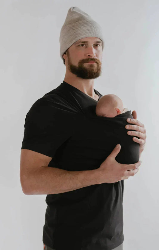 Mann mit schwarzem T-Shirt und Babytrage, graue Mütze, hält Baby sicher.