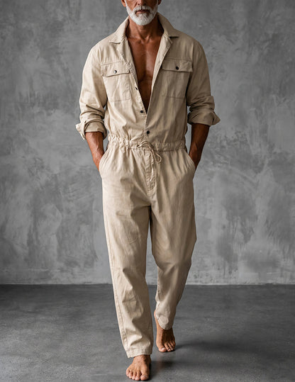 Man i beige jumpsuit med uppvikta ärmar och händerna i fickorna, barfota framför en grå betongvägg. 1