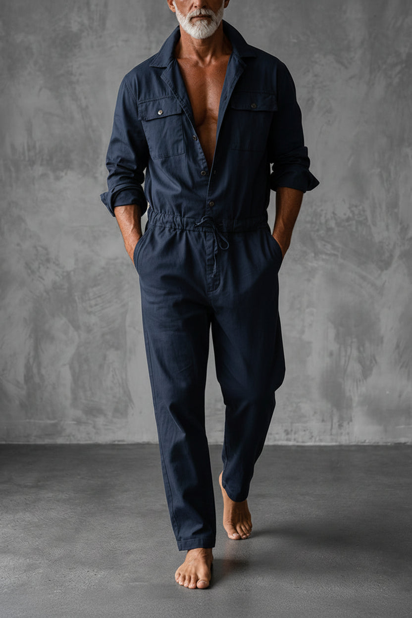 Man i marinblå jumpsuit med bröstfickor och dragsko i midjan, barfota mot en grå väggbakgrund. 2