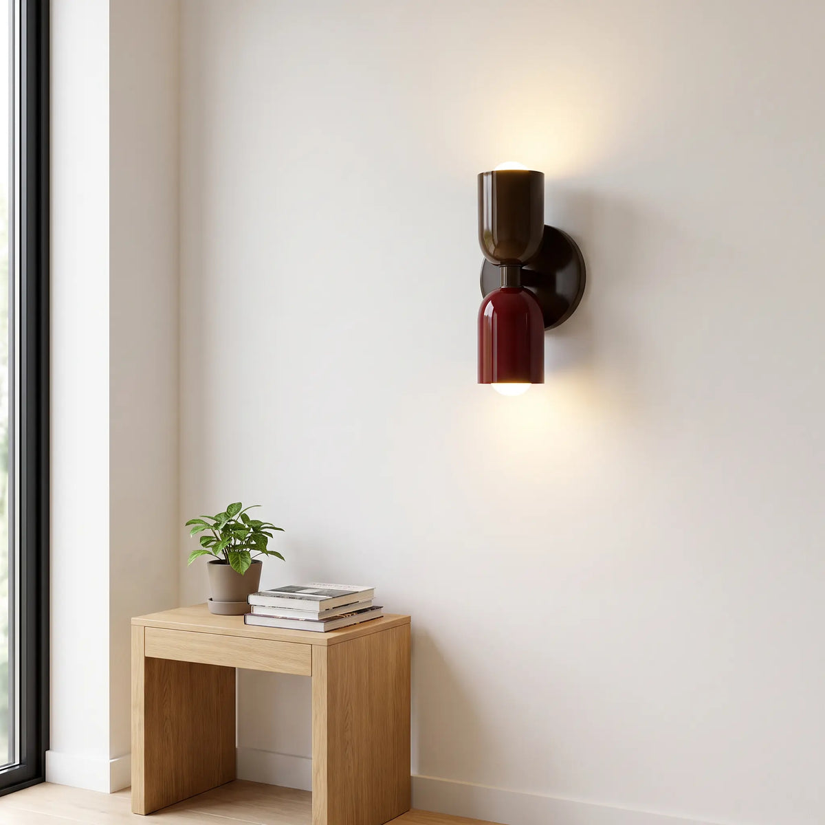 Vägglampa i mörkbrunt och vinrött tänd mot en vit vägg, med litet träbord, böcker och krukväxt nedanför. 10