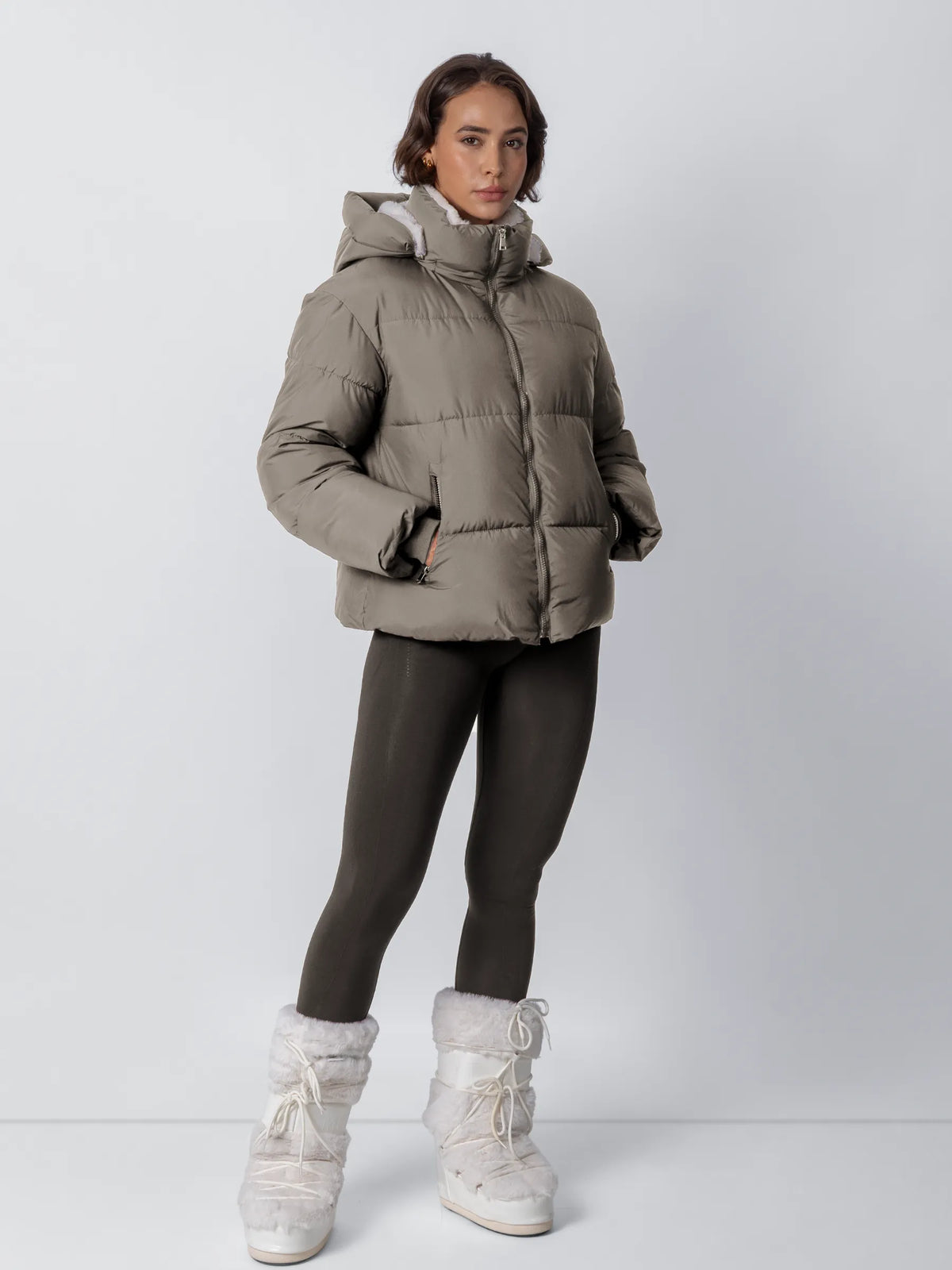 Modell i helfigur bär den gråbruna vadderade jackan med huva, mörka leggings och vita pälsskor 21