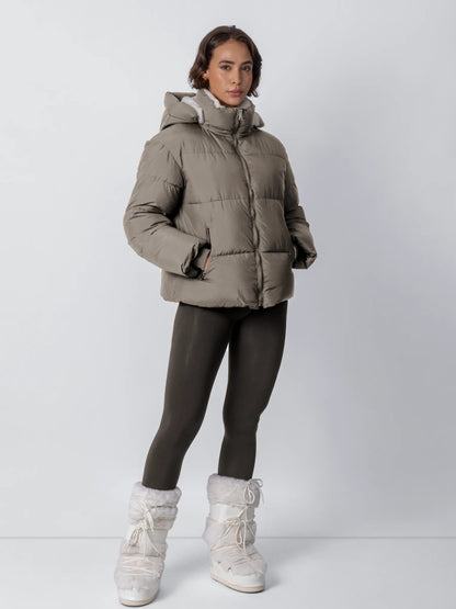 Modell i helfigur bär den gråbruna vadderade jackan med huva, mörka leggings och vita pälsskor 21