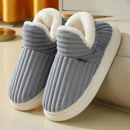 Ett par ribbade plyschtofflor i gråblått med vitt fluffigt foder och tjock vit sula, lutade mot en beige soffa. 2