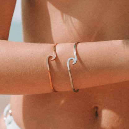 Zwei Wellenarmbänder aus Metall in Gold und Silber am Arm einer Person am Strand.