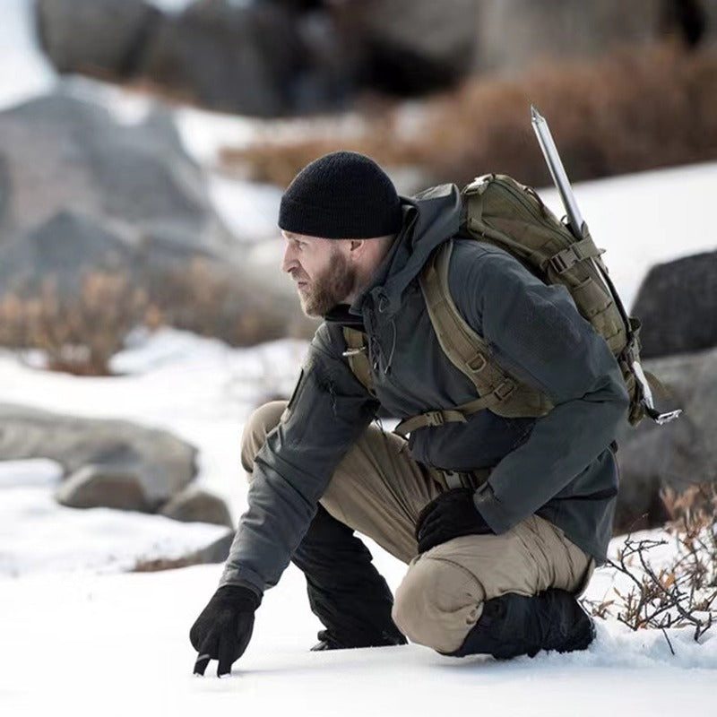 Mann in taktischer Winterkleidung mit Rucksack und Mütze im Schnee, Outdoor-Abenteuer.