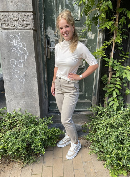 Frau in weißem Rollkragen-T-Shirt und grauer Hose vor grüner Tür, lächelnd, draußen.