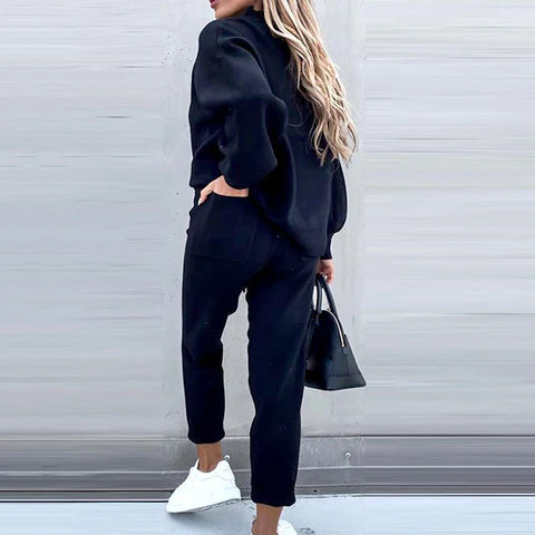 Frau in schwarzem Hausanzug, zweiteilig, mit Rollkragen und High-Waist-Hose, trägt weiße Sneaker.