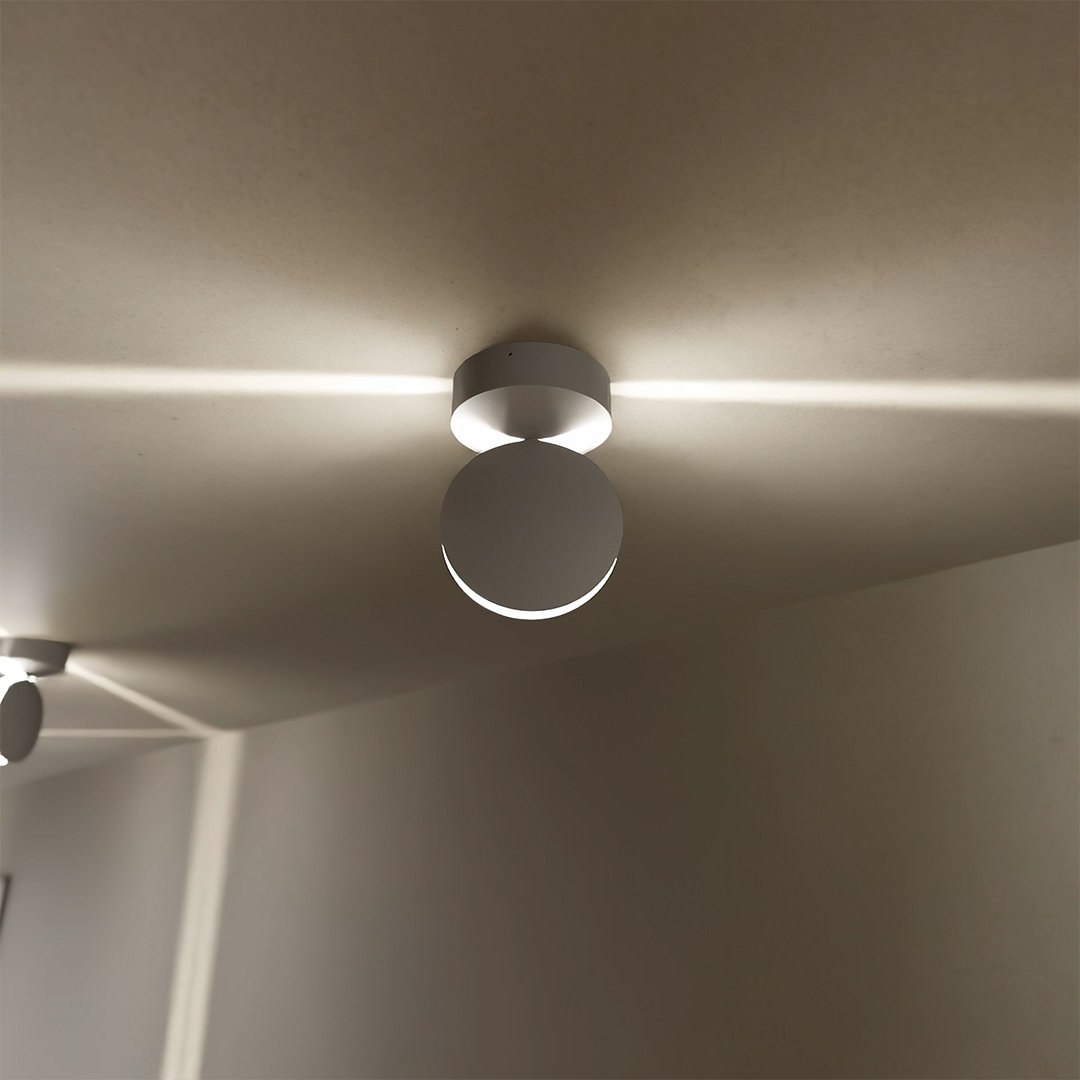 Närbild underifrån på en vit takmonterad lampa med rund rörlig skiva som sprider ljus åt båda sidor längs taket. 3