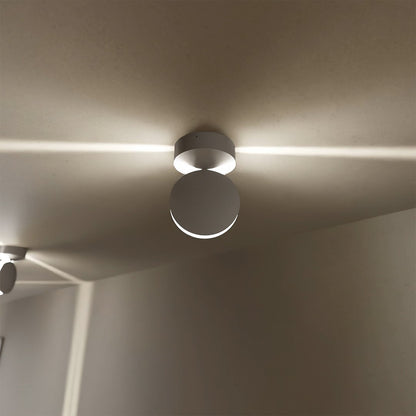 Närbild underifrån på en vit takmonterad lampa med rund rörlig skiva som sprider ljus åt båda sidor längs taket. 3