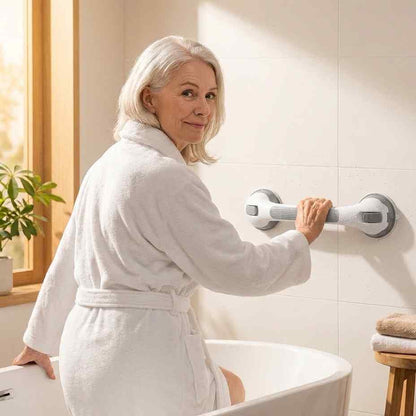 Äldre kvinna i vit badrock sitter på badkarskanten och håller i det vitgrå greppet som sitter fast på en kaklad vägg. 1